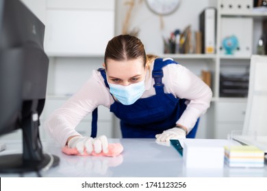 Woman In Face Mask, Gloves And Blue Overall Disinfecting Office To Prevent Spread Of Coronavirus, Cleaning Desk