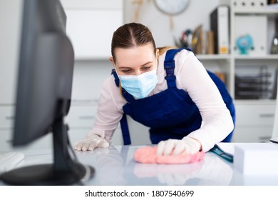 Woman In Face Mask, Gloves And Blue Overall Disinfecting Office To Prevent Spread Of Coronavirus, Cleaning Desk