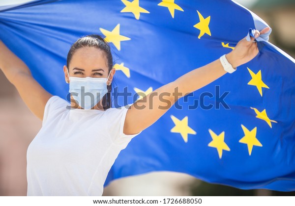 Woman in a face mask holding an European union flag behind her back with arms up.