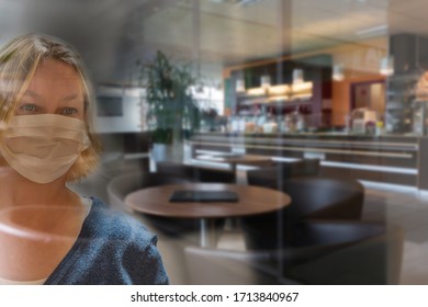 Woman With Face Mask Is Looking Throught A Glass Panel Into An Empty Closed Restaurant, Lockdown, Shutdown In Corona Crisis, Covid Pandemic