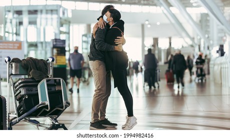 Woman In Face Mask Receiving Man At Airport Arrival. Couple Meeting And Hugging Each Other At Airport Arrival Gate.