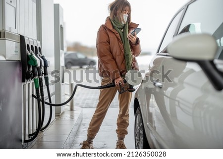 Woman in face mask refueling car with a gasoline, using smartphone to pay. Concept of mobile technology for fast refueling without visiting the store