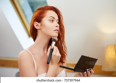 Woman face painting. Girl applying rouge or bronzing powder with brush to her skin in bathroom. Makeup cosmetics and beauty procedures.