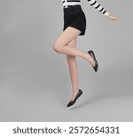 Woman with a fair complexion in a striped black-and-white sweater and high-waisted black shorts, playfully jumping while wearing black textured ballet flats, exuding a modern French-inspired flair.