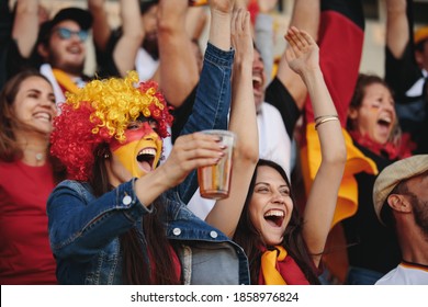 Woman In Fan Zone With A Wig And Face Painted In German Flag Colors Holding A Glass Of Beer And Cheering With Crowd Of Spectators At Sports Event.