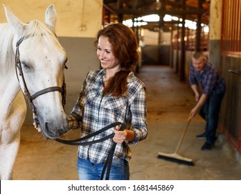 Woman farmer standing with white horse, man cleaning floor at stabling 
