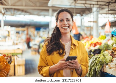 Woman at the farmer's market shopping, sending a text message on her smart phone. Young cheerful woman at the market. Smiling girl decided to cook a delicious and healthy meal - Powered by Shutterstock