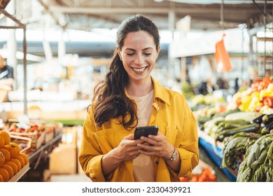 Woman at the farmer's market shopping, sending a text message on her smart phone. Young cheerful woman at the market. Smiling girl decided to cook a delicious and healthy meal - Powered by Shutterstock