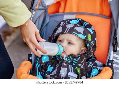 Woman Is Feeding Baby In Pram From Bottle Outside.