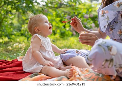 Woman Feeding Child With Spoon. Mom Feed Baby With Pureed Food. Mom And Daughter In A Dress Spend Their Leisure Time In The Park In The Summer