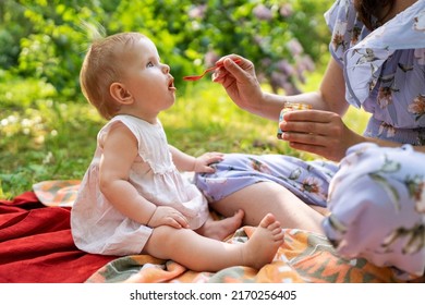 Woman Feeding Child With Spoon. Mom Feed Baby With Pureed Food. Mom And Daughter In A Dress Spend Their Leisure Time In The Park In The Summer