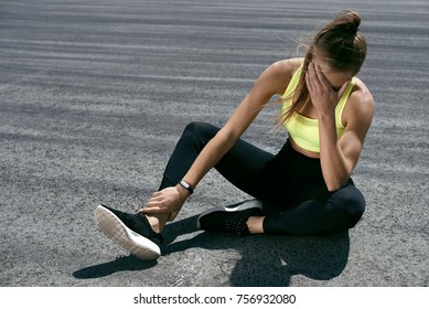 Woman Feeling Tired After Running Outdoors. Sad Exhausted Disappointed Female Athlete In Sportswear Sitting On Ground After Losing Sports Competition, Feeling Pain In Injured Leg. High Resolution.