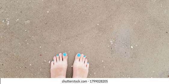 Woman Feet With Blue, Pink Toenails On Natural Beach Sand And Seashells.