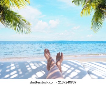 Woman Feet On Sunbath Chair With Coconut Leaves Shadow Over Summer Beach With Blue Sky And White Clouds Landscape Background.