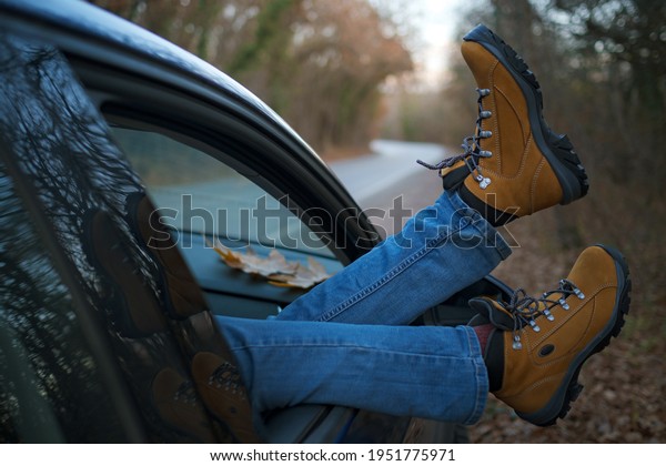 Woman feet in trendy yellow\
hiking boots on car door. Feet outside the window at sunset forest.\
The concept of freedom of movement. An autumn weekend in\
nature.