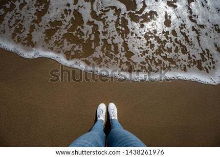 Woman feet view with jeans from above at the beach with sand and wave coming in the frame with copy space