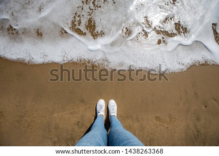 Woman feet view with jeans from above at the beach with sand and wave coming in the frame with copy space