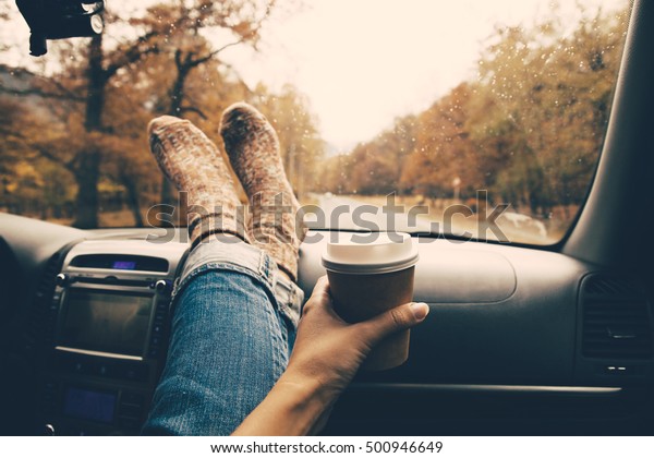 Woman feet in warm socks on car\
dashboard. Drinking take away coffee on road. Fall trip. Rain drops\
on windshield. Freedom travel concept. Autumn\
weekend.