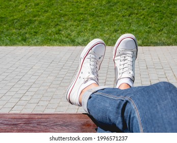 Woman feet in white sneakers on grass lawn background. Female has a rest on wooden bench in urban park at sunny day. Summer vibes. Slow living. Enjoying sunlight and peace outdoors.