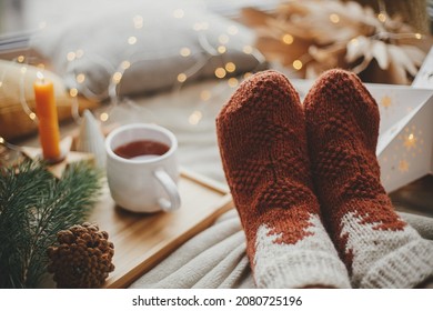 Woman feet in woolen socks on soft bed with warm cup of tea, christmas stars, golden lights, trees, candle and pillows. Cozy winter moments at home. Girl relaxing in scandinavian evening room - Powered by Shutterstock