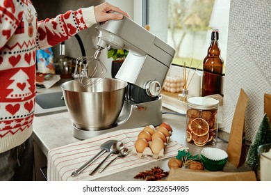 Woman in festive christmas sweater preparing dough for cookies at home kitchen. Female hands use electric mixer. Modern kitchen household appliances - Powered by Shutterstock