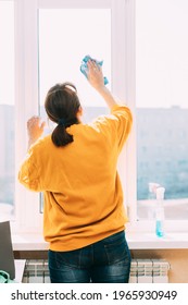 Woman Of Fifty In Yellow Sweater And Jeans Washes Dusty Window In Apartment. 50 Year Old Woman Cleans Windows From Stains Using Rag And Spray Cleaner. Caucasian Elderly Woman Is Cleaning House, Doing