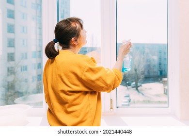 Woman Of Fifty In Yellow Sweater And Jeans Washes Dusty Window In Apartment. 50 Year Old Woman Cleans Windows From Stains Using Rag And Spray Cleaner. Caucasian Elderly Woman Is Cleaning House, Doing