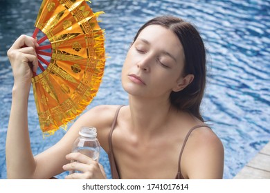 Woman Fighting Heat Wave With A Fan. Portrait Of A Young Woman Next To The Water During A Hot Summer Day.