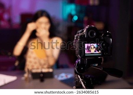 Woman filming makeup tutorial while being filmed by a professional camera in studio, with blurred purple neon background. Female influencer making content for vlog