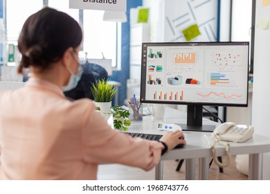 Woman In Financial Company Wearing Face Mask Looking At Statistics, Charts, As Prevention During Global Outbreak With Covid19 Flu. Safety Shiled Between Coworkers.