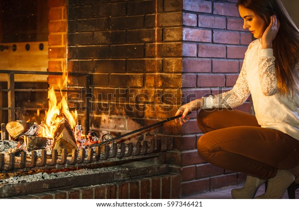 Woman with fire iron poker\
at fireplace. Young girl heating warming up and relaxing. Winter at\
home.