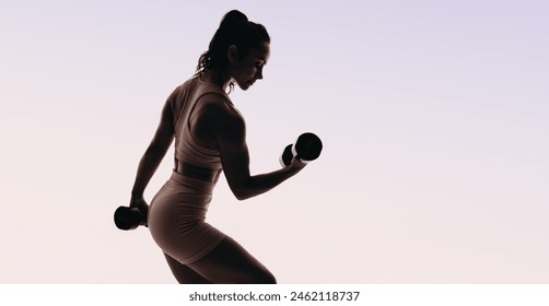 Woman in fitness clothing engages in weight training and bodybuilding, demonstrating form and muscular strength. Athletic woman lifting dumbbells in a studio, silhouetted against a pink background. - Powered by Shutterstock