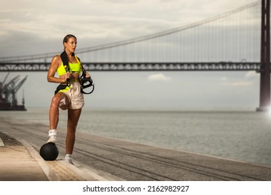Woman in fitness wear doing workout with a heavy medicine ball and trx straps outdoors.. Sporty gitl with fit body in sportswear posing on sky background.