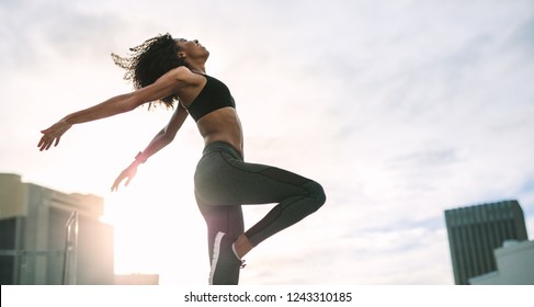 Woman in fitness wear jumping high standing on rooftop with sun flare in the background. Female athlete doing exercises in the morning on terrace of a building. - Powered by Shutterstock