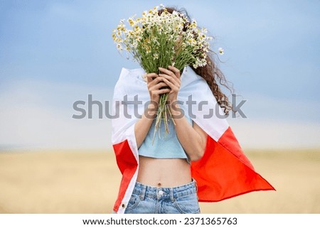 Woman with flag of Poland on shoulders holding bouquet of daisies in front of face in wheat field. Polish Flag Day. Independence Day. Travel and learn polish language concept. Selective focus.