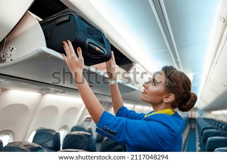 Woman flight attendant or air hostess placing travel bag in overhead baggage locker while standing in airplane passenger salon