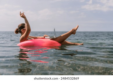 A woman is floating on a pink inflatable raft in the ocean while using a laptop. Concept of relaxation and leisure, as the woman enjoys her time in the water while working on her laptop. - Powered by Shutterstock