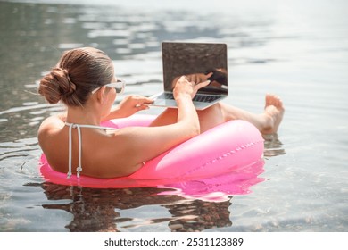 A woman is floating on a pink inflatable raft in the ocean while using a laptop. Concept of relaxation and leisure, as the woman enjoys her time in the water while working on her laptop. - Powered by Shutterstock