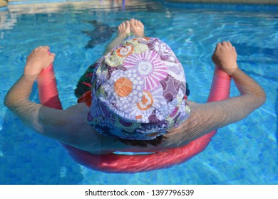 Woman Floating On A Pool Noodle In A Swimming Pool, View From Behind Wearing A Hat