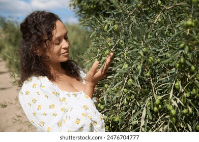 A woman in a floral dress examines an olive branch in an orchard. She is surrounded by vibrant, green olive trees under a clear, sunny sky. - Powered by Shutterstock