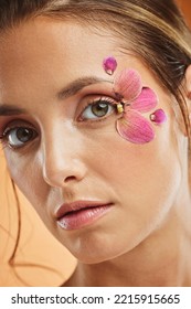 Woman, Flower And Beauty Eye Makeup In Portrait For Cosmetics, Skincare And Facial Wellness Mockup In Studio Against Orange Background. Model, Cosmetic Art And Orchid Petals Design For Spring