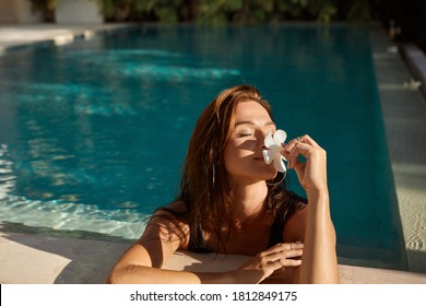 Woman with flower in her hand relaxing in outdoor swimming pool in Bali luxury resort. Beautiful young woman smell  flowers at swimming pool