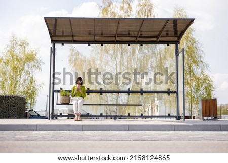 Woman with flowerpot at modern bus stop outdoors, wide front view with copy space. Concept of public transportation and sustainability