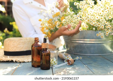 Woman With Flowers Near Table Outdoors, Focus On Bottles Of Chamomile Essential Oil