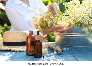 Woman With Flowers Near Table Outdoors, Focus On Bottles Of Chamomile Essential Oil