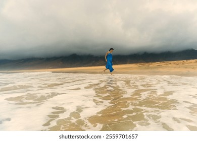 A woman in a flowing blue dress running barefoot on the sandy Playa de Cofete beach in Fuerteventura, Canary Islands. The dramatic cloudy sky, golden sand, and ocean waves  - Powered by Shutterstock