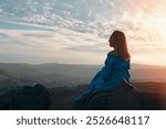 A woman in a flowing blue gown gazes at the sunset over a vast, serene landscape of rolling hills and valleys