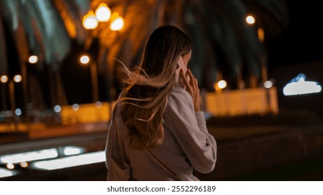 A woman with flowing hair speaks on a phone at night, illuminated by warm streetlights, with palm trees and a blurred cityscape in the background. - Powered by Shutterstock