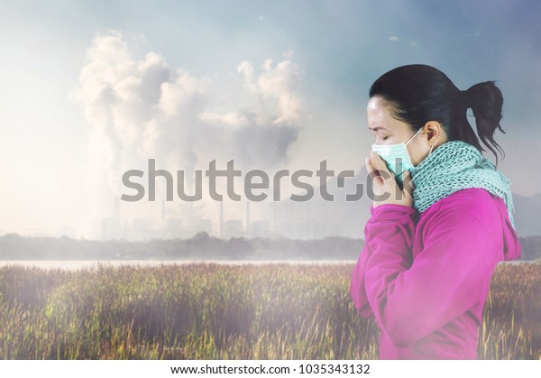 Woman with flu sneezing, woman\
dressed pink in winter clothing wearing mask to her nose in a cold\
and flu health concept against air pollution\
background.