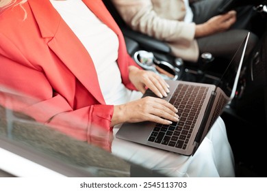 A woman is focused on her laptop while seated in a car. The interior is modern and stylish. Outside, city life continues with blurred movements visible through the window. - Powered by Shutterstock